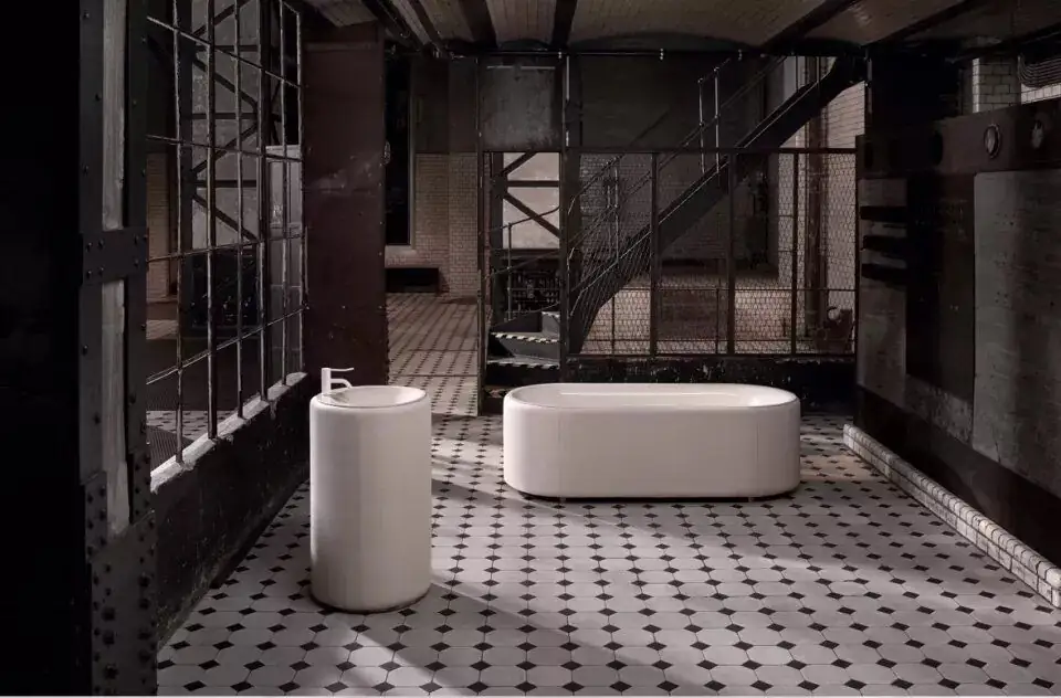 Bañera ovalada exenta y lavabo de pie cilíndrico tapizados en tejido color crudo, en una nave industrial con suelo de baldosa octogonal blanca y negra