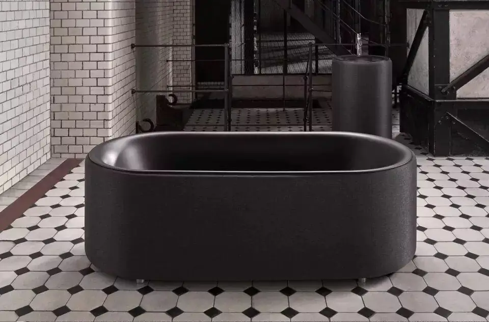 Bañera ovalada exenta tapizada en tejido gris antracita con interior oscuro y, al fondo, un lavabo de pie del mismo color
