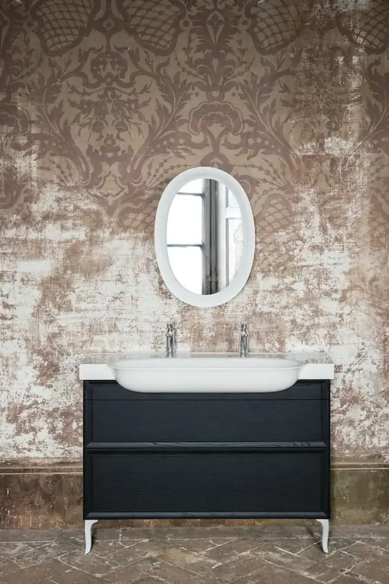 Lavabo blanco sobre un mueble negro de dos cajones con patas curvadas, bajo un espejo ovalado, frente a una pared con papel de damasco