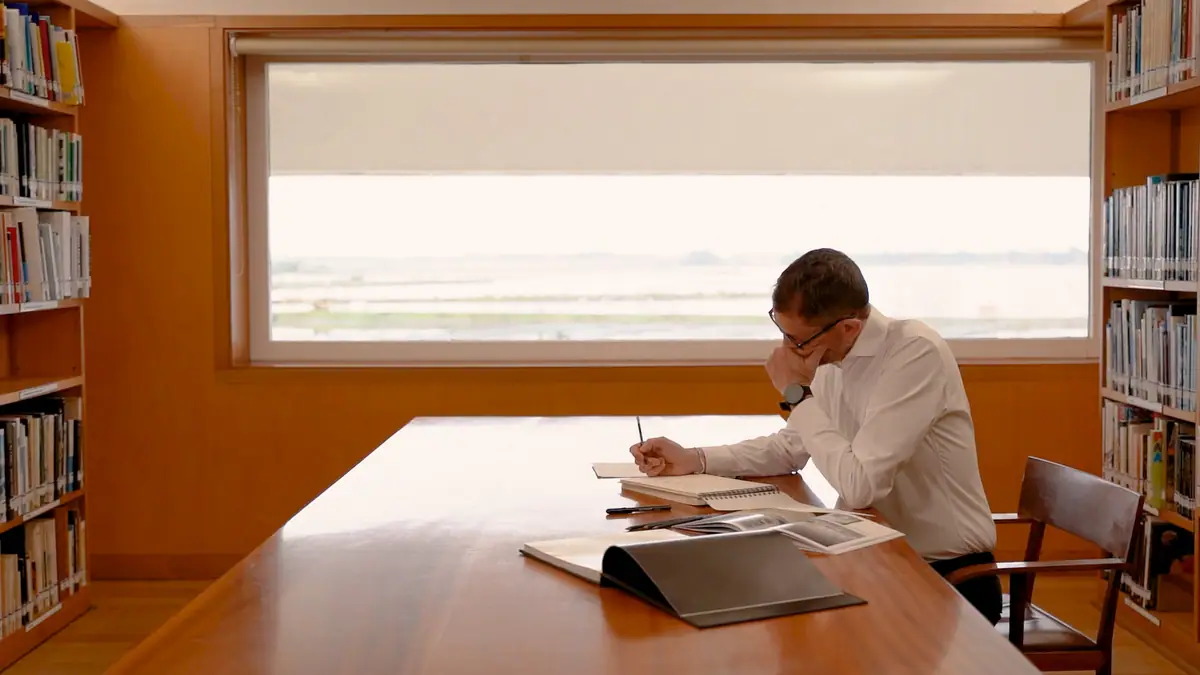 Hombre con camisa blanca dibujando en una mesa de madera de una biblioteca, ante un gran ventanal