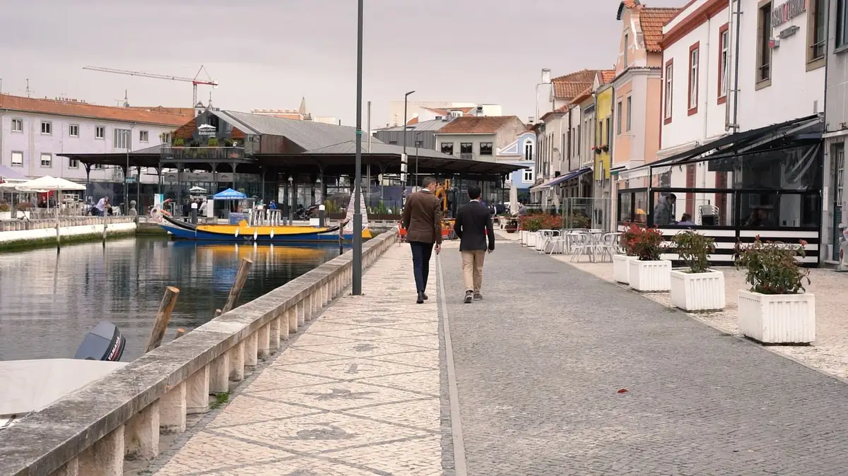 Dos hombres caminan por un paseo junto a un canal con un barco amarillo y azul y fachadas de colores