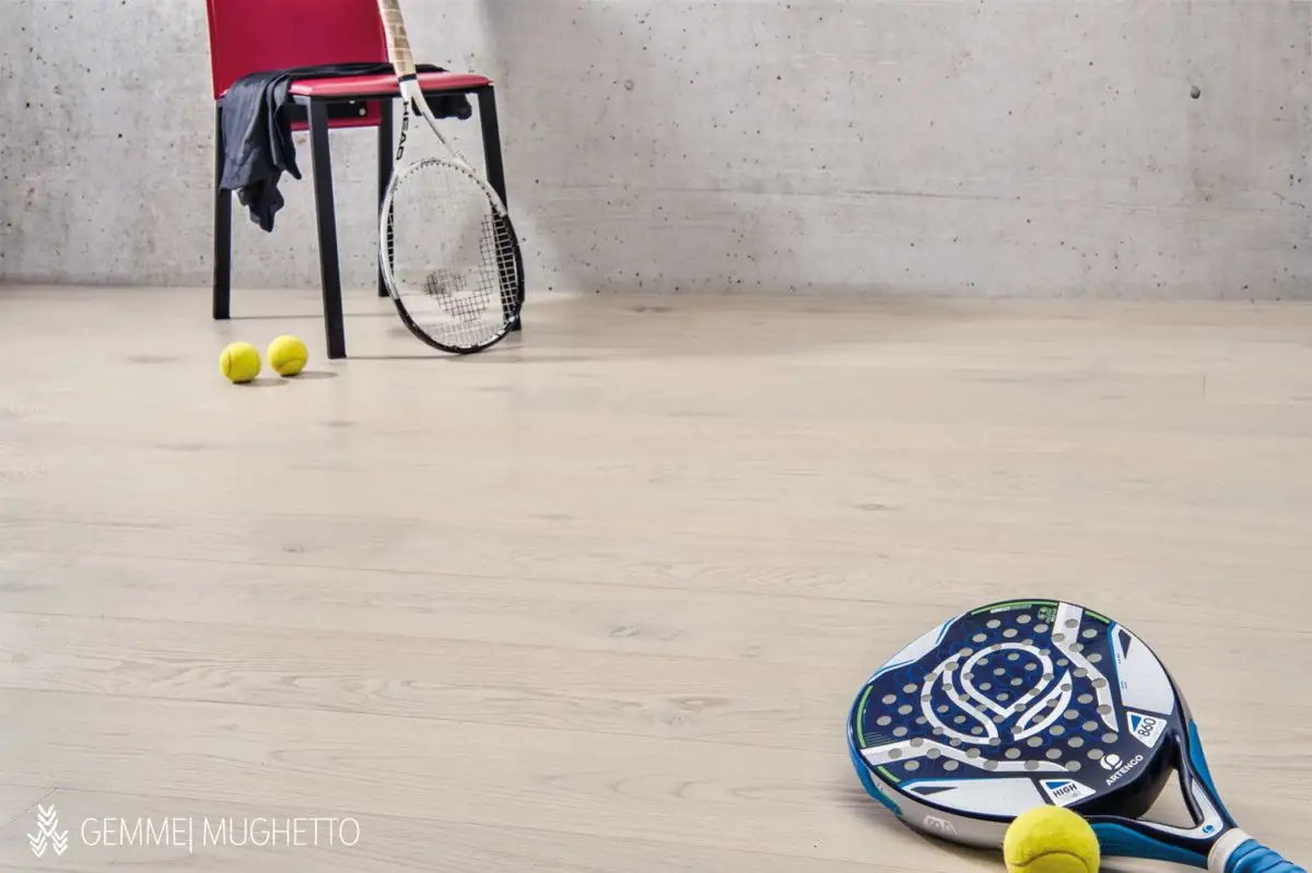 Suelo de tablas de roble muy claro con una silla roja, una raqueta de tenis, pelotas y una pala de pádel