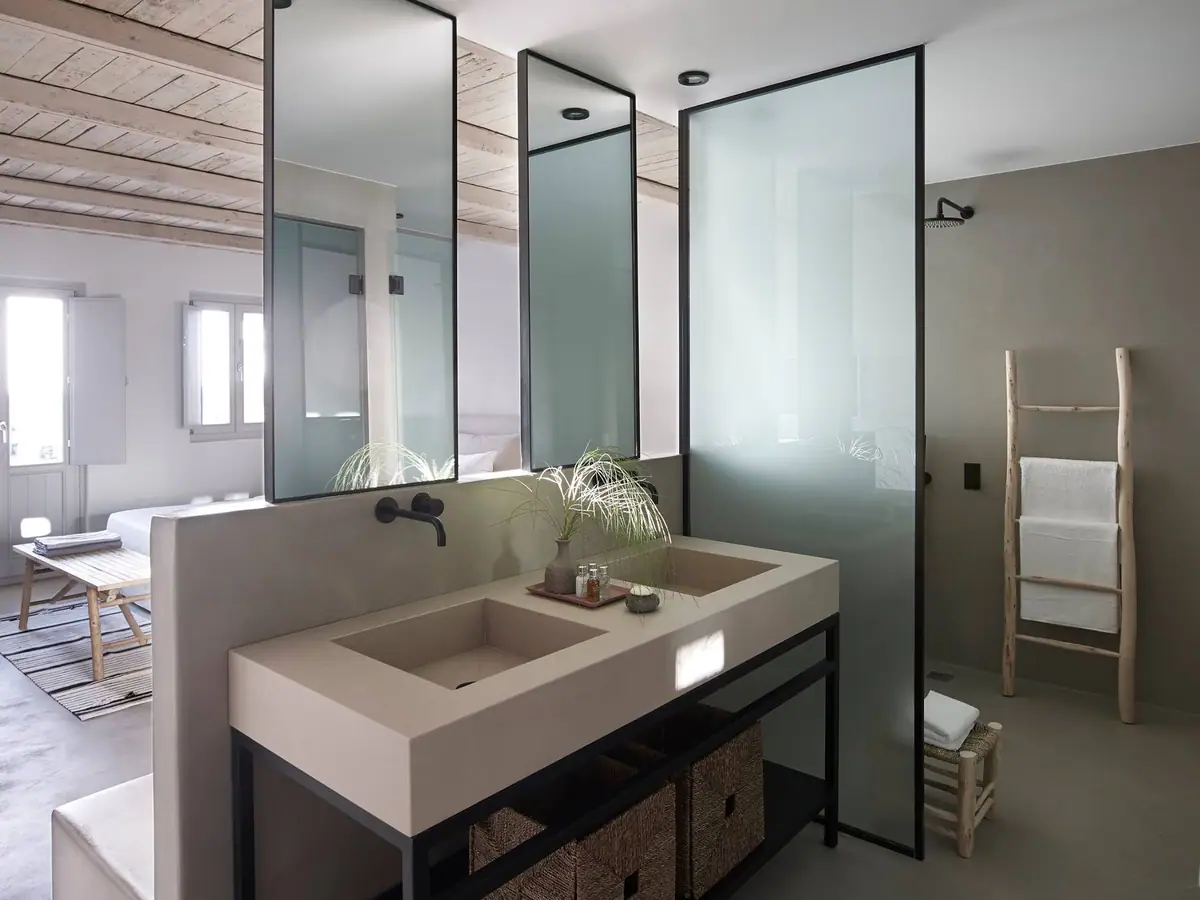 Baño de hotel con doble lavabo de obra en tono arena, grifería mural negra y mampara de vidrio translúcido.