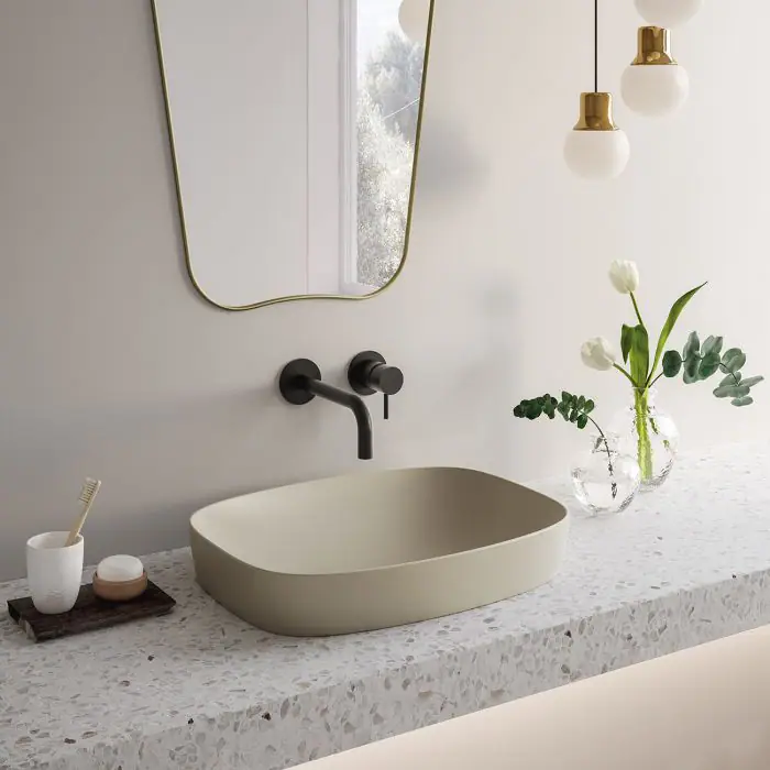 Lavabo de cerámica de líneas suaves sobre una encimera de terrazo, con grifería mural negra y un espejo de marco dorado