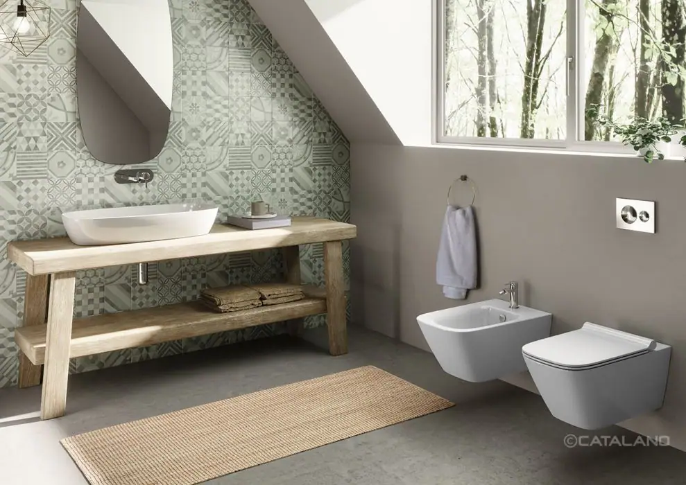 Baño abuhardillado con un lavabo blanco sobre un banco de madera, pared de baldosas hidráulicas verdes e inodoro y bidé suspendidos