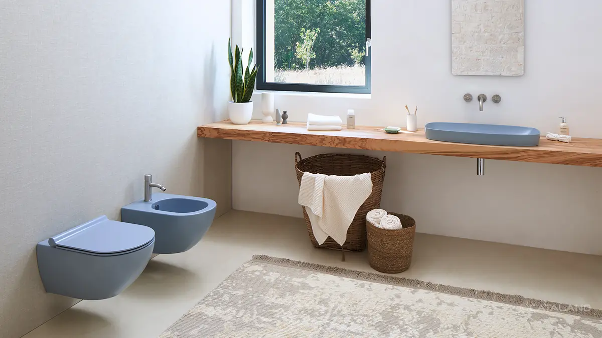 Baño con un lavabo azul satinado sobre una encimera de madera bajo la ventana y un inodoro y un bidé suspendidos del mismo color