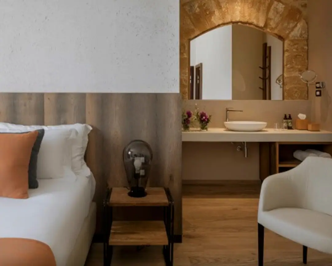 Dormitorio de hotel con zona de lavabo abierta, lavabo blanco de apoyo sobre encimera clara y espejo enmarcado por un arco de piedra