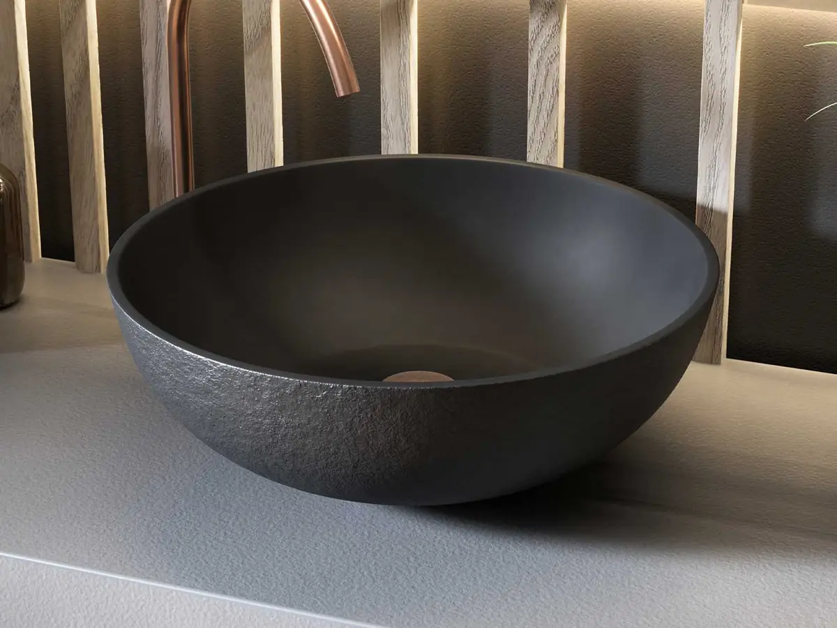 Lavabo redondo de apoyo en color oscuro con exterior texturizado, sobre encimera gris y con grifo de caño alto en cobre