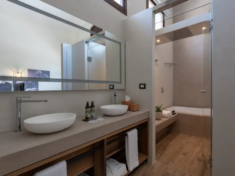 Baño de hotel con dos lavabos ovalados blancos sobre encimera clara con mueble de madera abierto y gran espejo