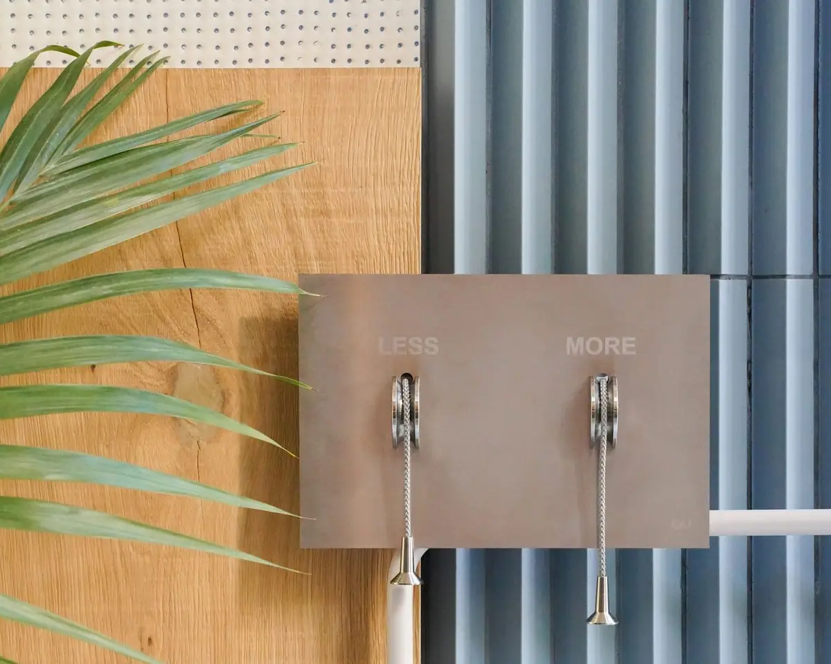 Pulsador LESS is MORE de acero inoxidable con dos cordones trenzados, sobre una pared de lamas azules junto a un panel de madera y una hoja de palmera