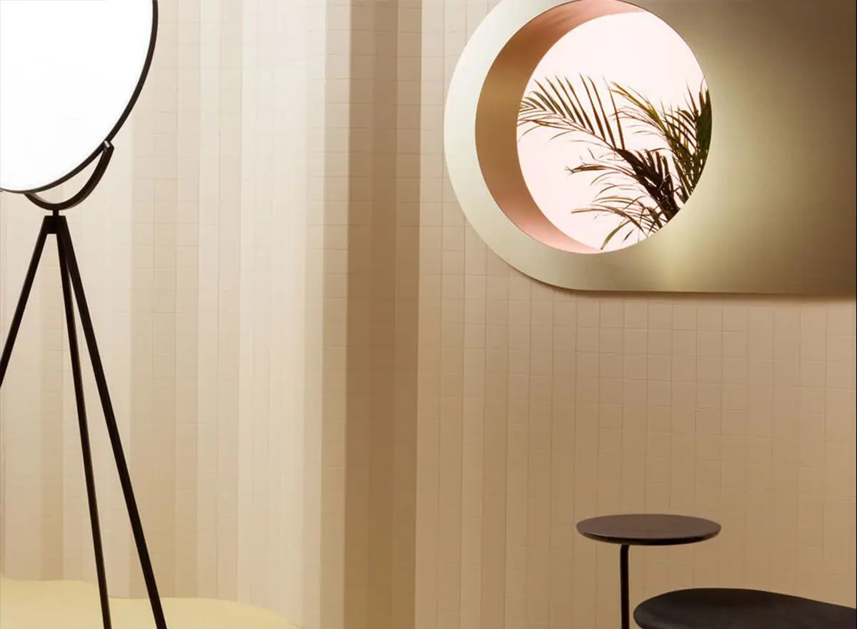 Pared revestida con una malla de piezas rectangulares en tono crema que forma pliegues verticales, junto a una lámpara de pie y un espejo redondo.