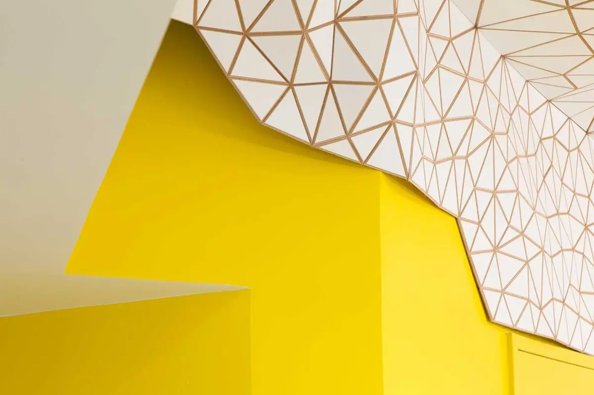Detalle del encuentro entre la malla teselada blanca del techo y una pared pintada de amarillo intenso.
