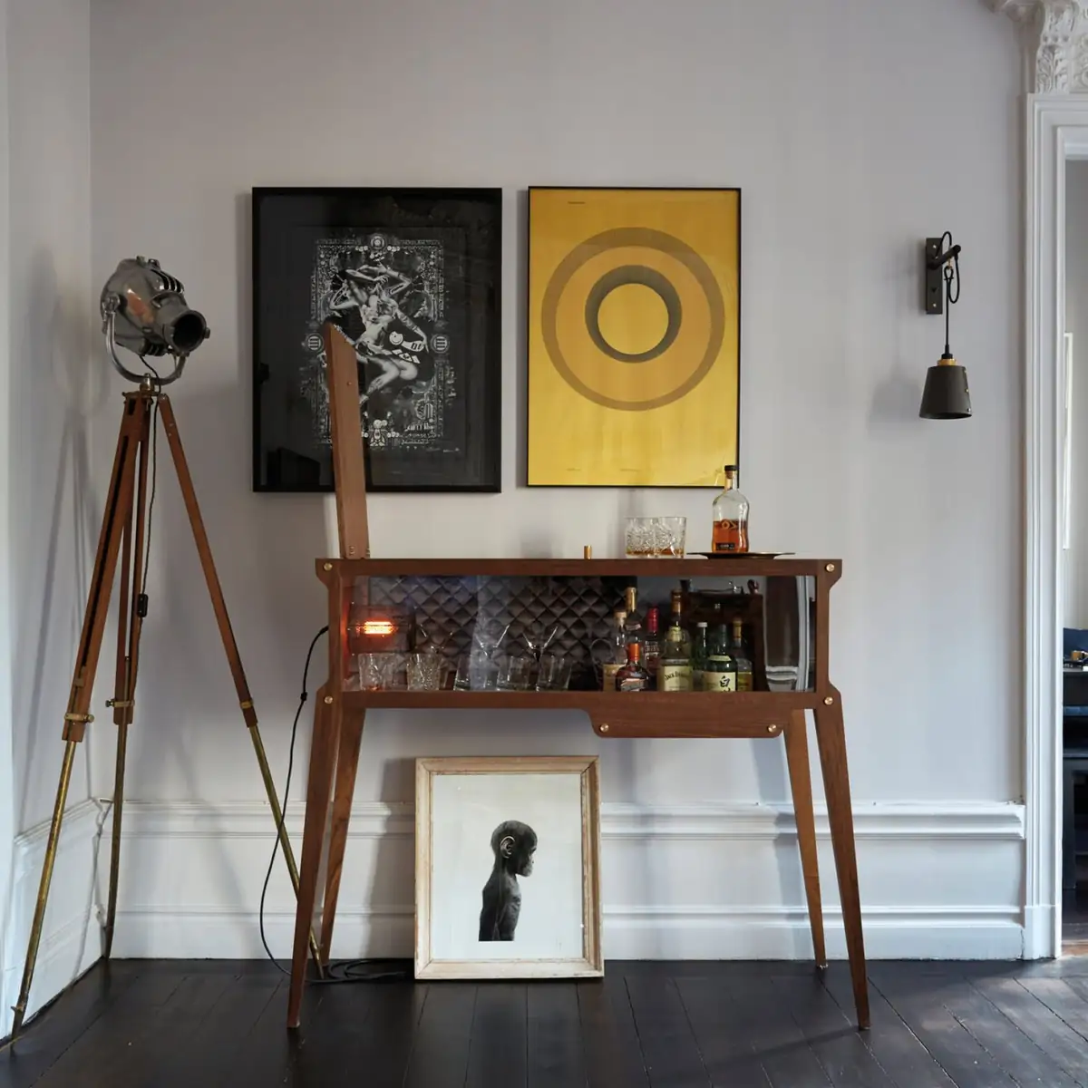 Mueble bar de madera con el panel trasero abierto, botellas y vasos iluminados, junto a un foco sobre trípode