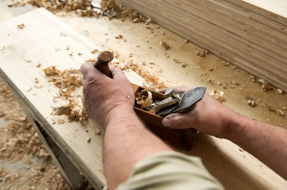 Manos de un artesano cepillando una tabla de madera con un cepillo de carpintero, entre virutas.