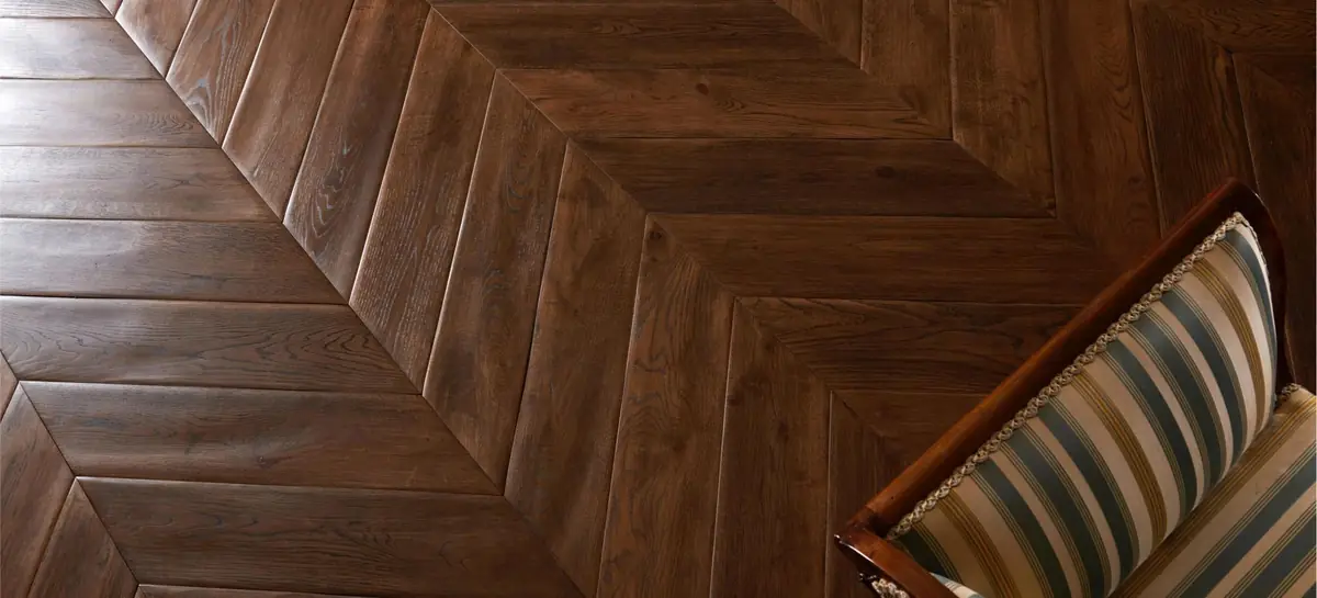 Pavimento de madera oscura en espiga húngara de piezas anchas junto a un sillón tapizado.