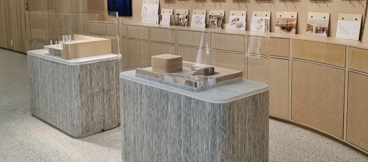 Dos peanas expositivas revestidas con chapa de ratán gris con maquetas sobre ellas, en un interior con mobiliario de madera.