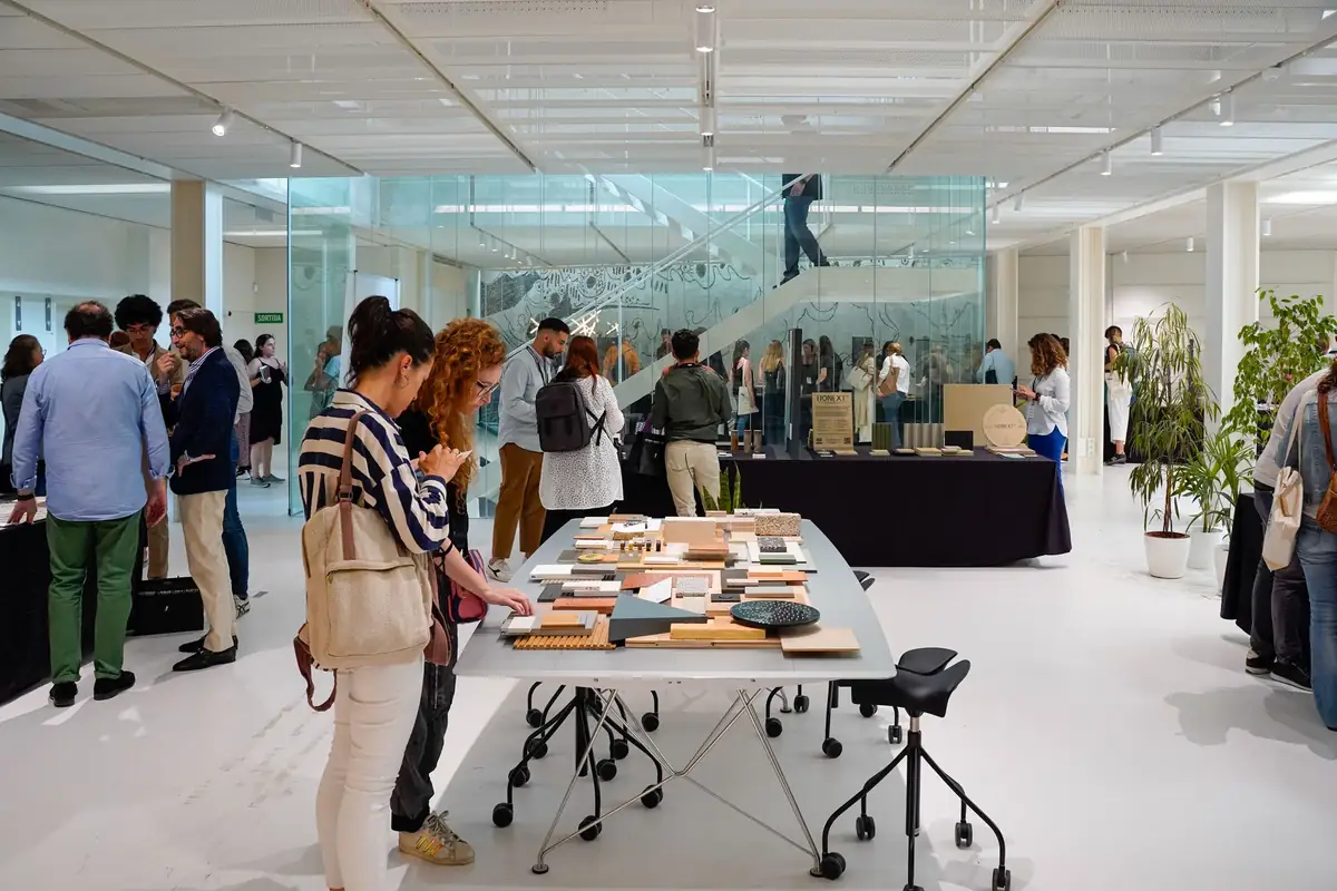 Visitantes alrededor de una gran mesa blanca con muestras de materiales en la sala de exposición del COAC.