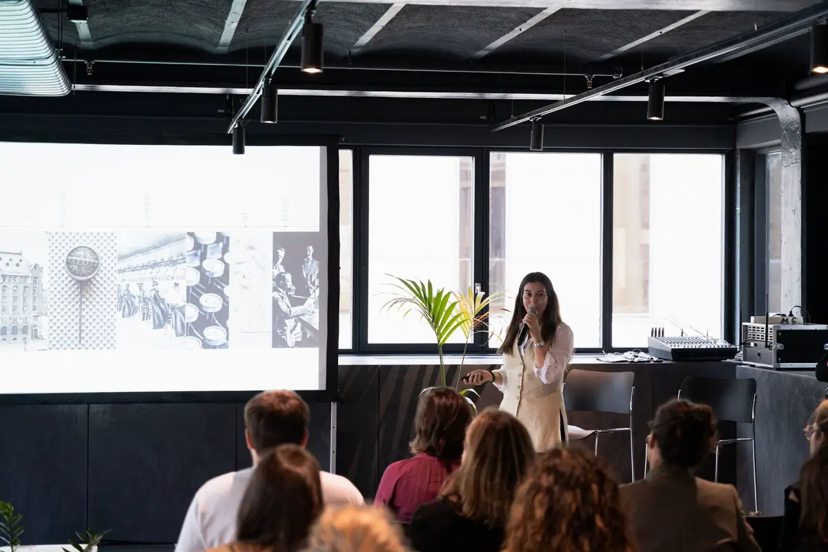 Ponente hablando al público junto a una proyección de imágenes de archivo de arquitectura.