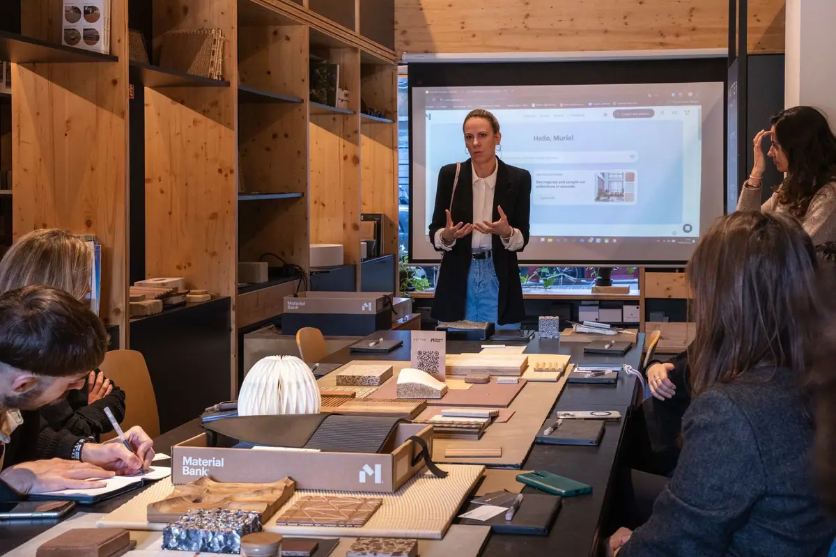 Presentación de la plataforma de Material Bank proyectada en pantalla ante los asistentes, con muestras y cajas de Material Bank sobre la mesa.