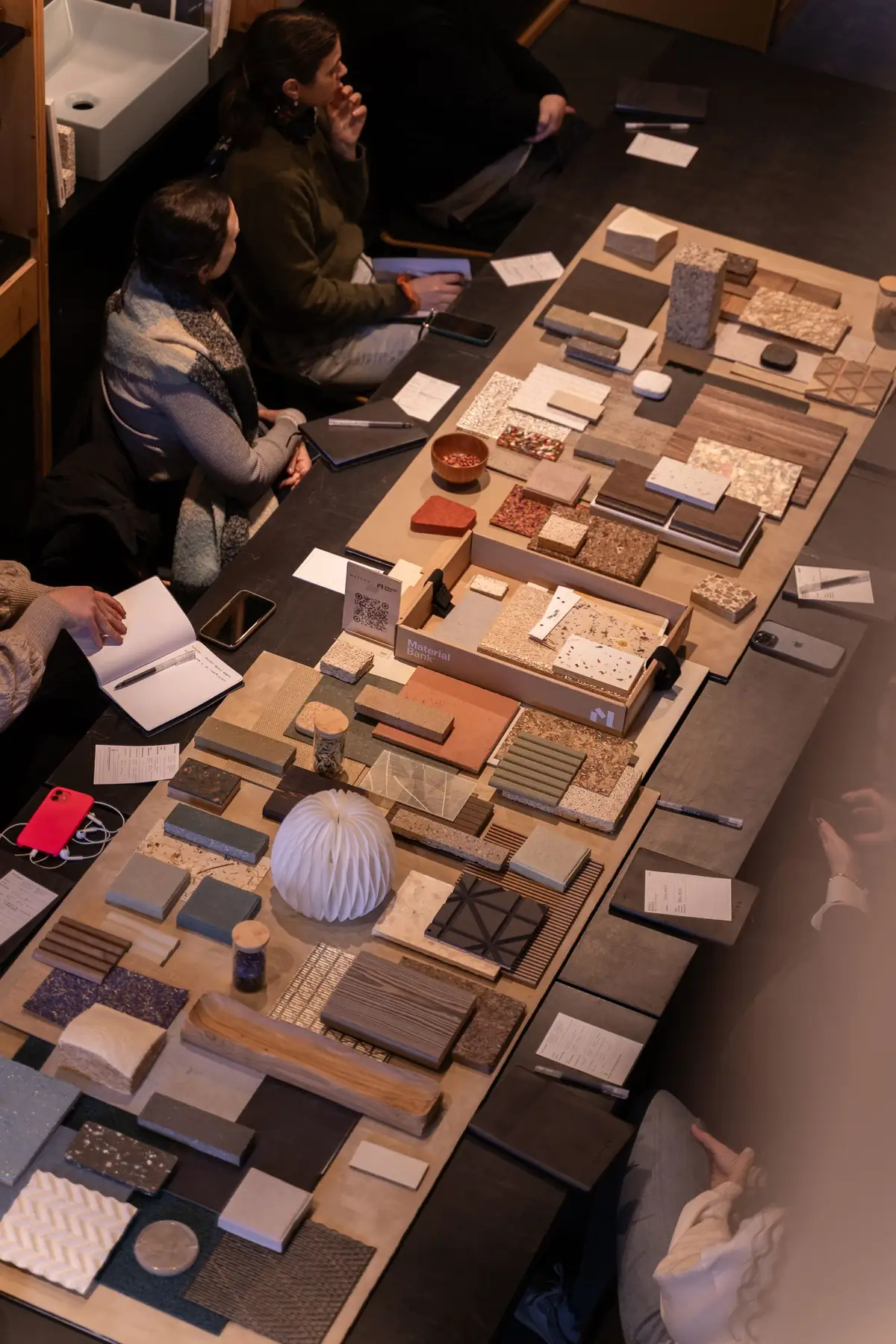 Vista cenital de una mesa con muestras de madera, aglomerados, terrazos, cerámica y textiles y una caja de Material Bank, mientras varias asistentes toman notas.