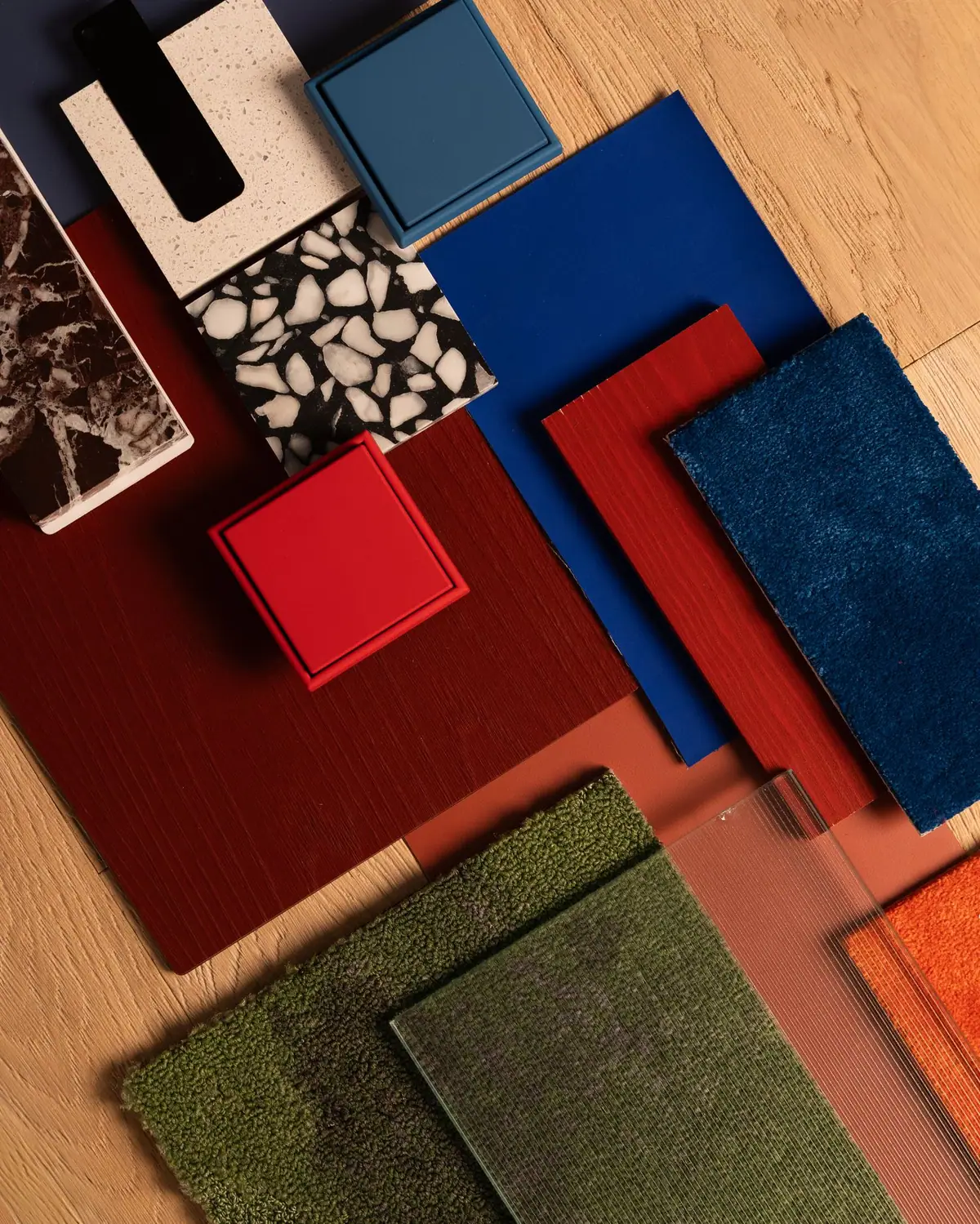 Composición de muestras de materiales en rojos, azules y verdes: moquetas, vidrio acanalado, terrazo y piedra sobre un suelo de madera
