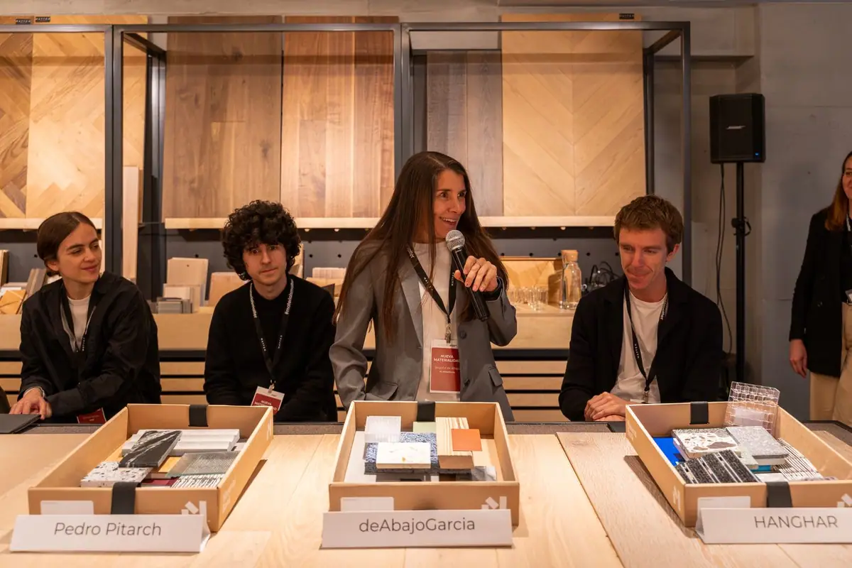 Una arquitecta habla con micrófono sentada a una mesa de madera con cajas de muestras rotuladas con el nombre de cada estudio