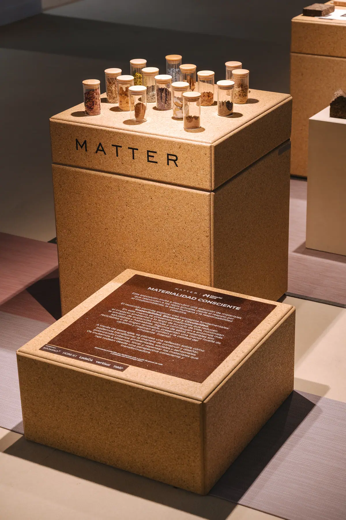 Peana con la marca Matter y frascos de materias primas, delante de un panel marrón con el texto de presentación de la exposición