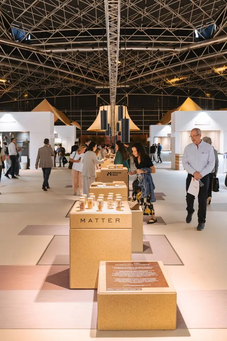 Visitantes recorren una exposición de materiales con peanas bajas bajo una cubierta de estructura metálica