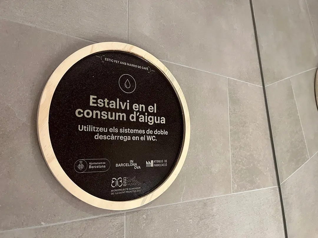 Señal circular marrón oscuro con un marco de madera clara y el texto en catalán Estalvi en el consum d'aigua, sobre una pared de baldosas grises