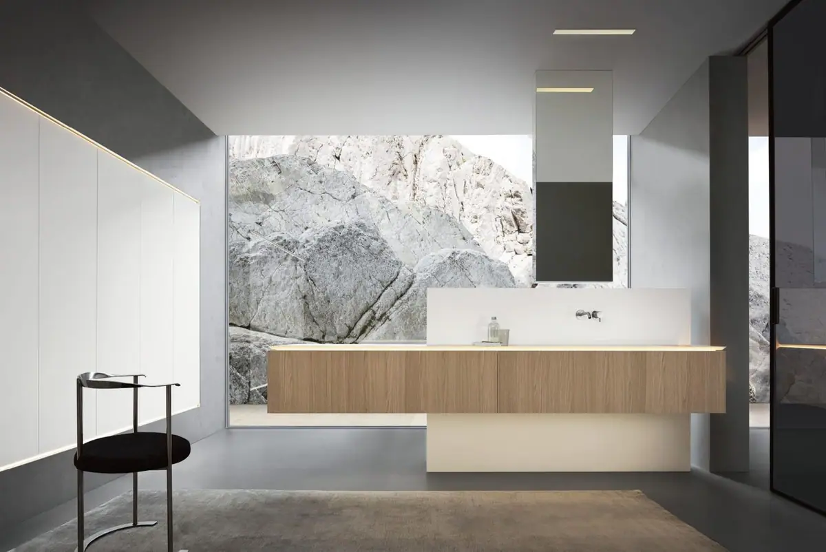 Baño con mueble suspendido de roble claro y encimera blanca iluminada por debajo, espejo colgado del techo y una gran cristalera abierta a unas rocas.