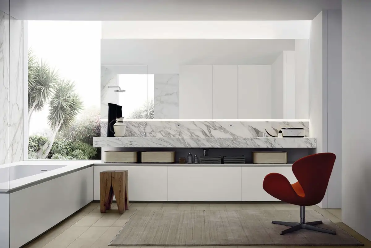 Baño blanco con una larga encimera de mármol Arabescato, bañera revestida en blanco, armarios altos y un sillón rojo junto a la ventana.