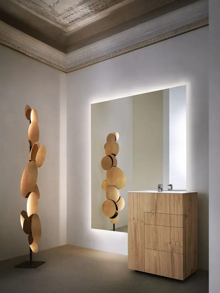 Mueble lavabo de pie de madera de roble con cajones, delante de un gran espejo retroiluminado, junto a una lámpara escultórica de discos de madera.