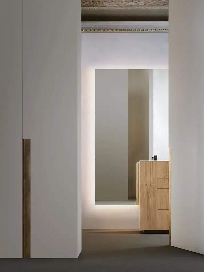 Vista lejana de un mueble lavabo de roble al fondo de un pasillo blanco, con un espejo alto retroiluminado.