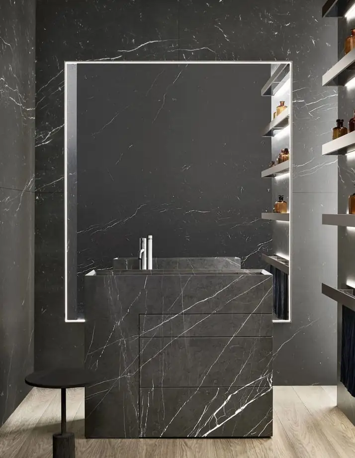 Bloque de lavabo de mármol negro con vetas blancas frente a una pared del mismo mármol, con espejo retroiluminado y estantes iluminados.