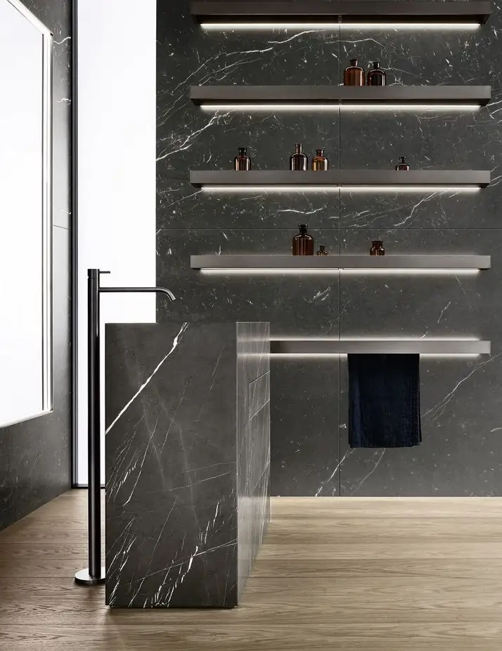 Lavabo monolítico de mármol negro visto de perfil, con un grifo alto de pie y estantes iluminados en la pared.