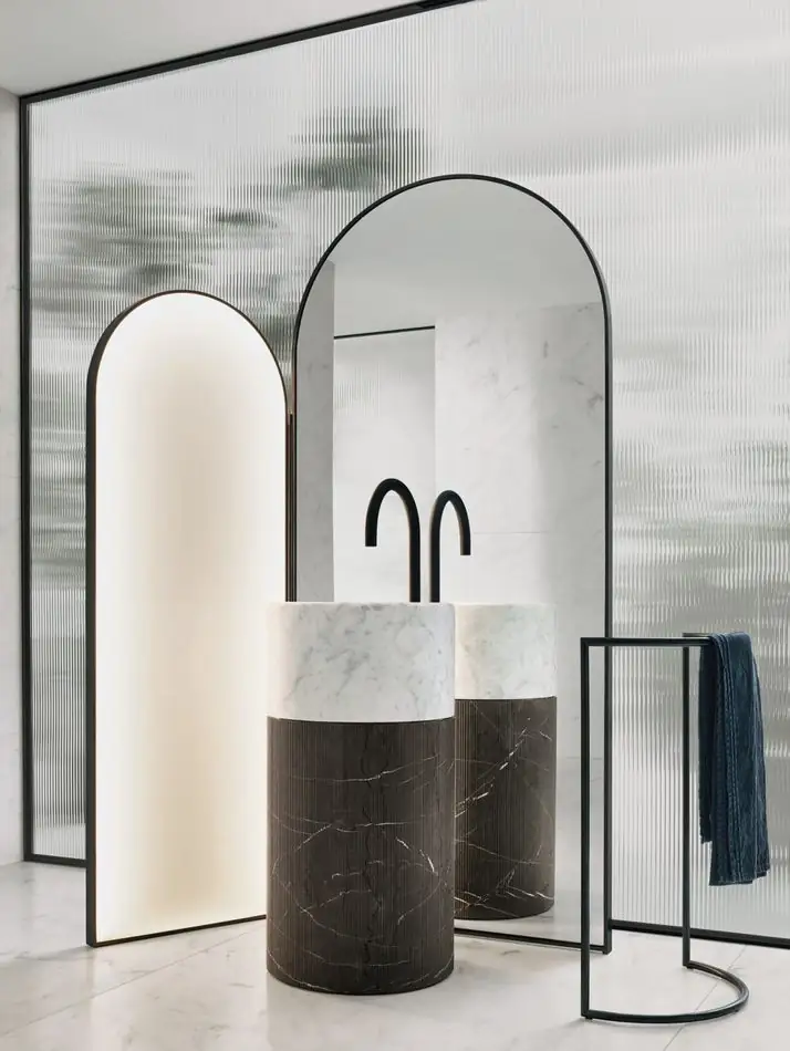 Lavabo cilíndrico de mármol blanco con base de mármol oscuro y grifo negro, entre espejos exentos de perfil negro y una pantalla de vidrio texturado.