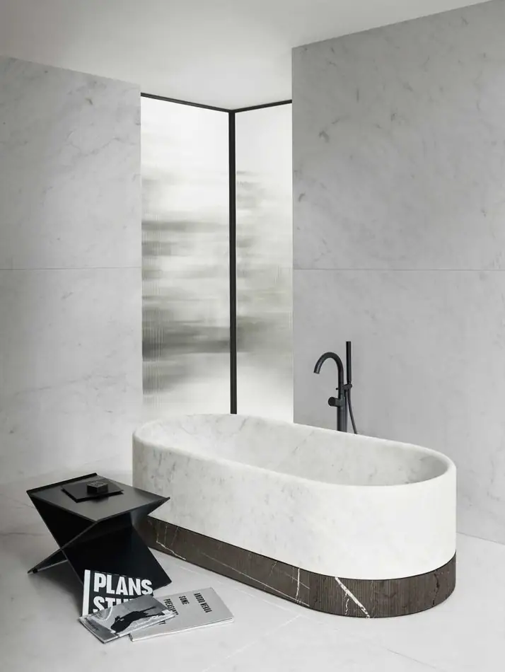 Bañera de mármol blanco de forma redondeada con zócalo de mármol oscuro y grifería negra de pie, en un baño de mármol claro.