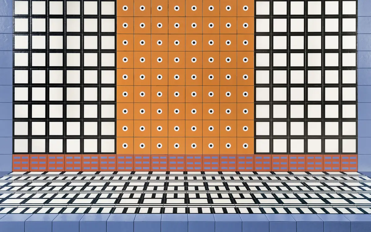 Pared de baldosas con cuadrados blancos enmarcados en negro, un paño central naranja con círculos y laterales azul claro, sobre suelo a rayas