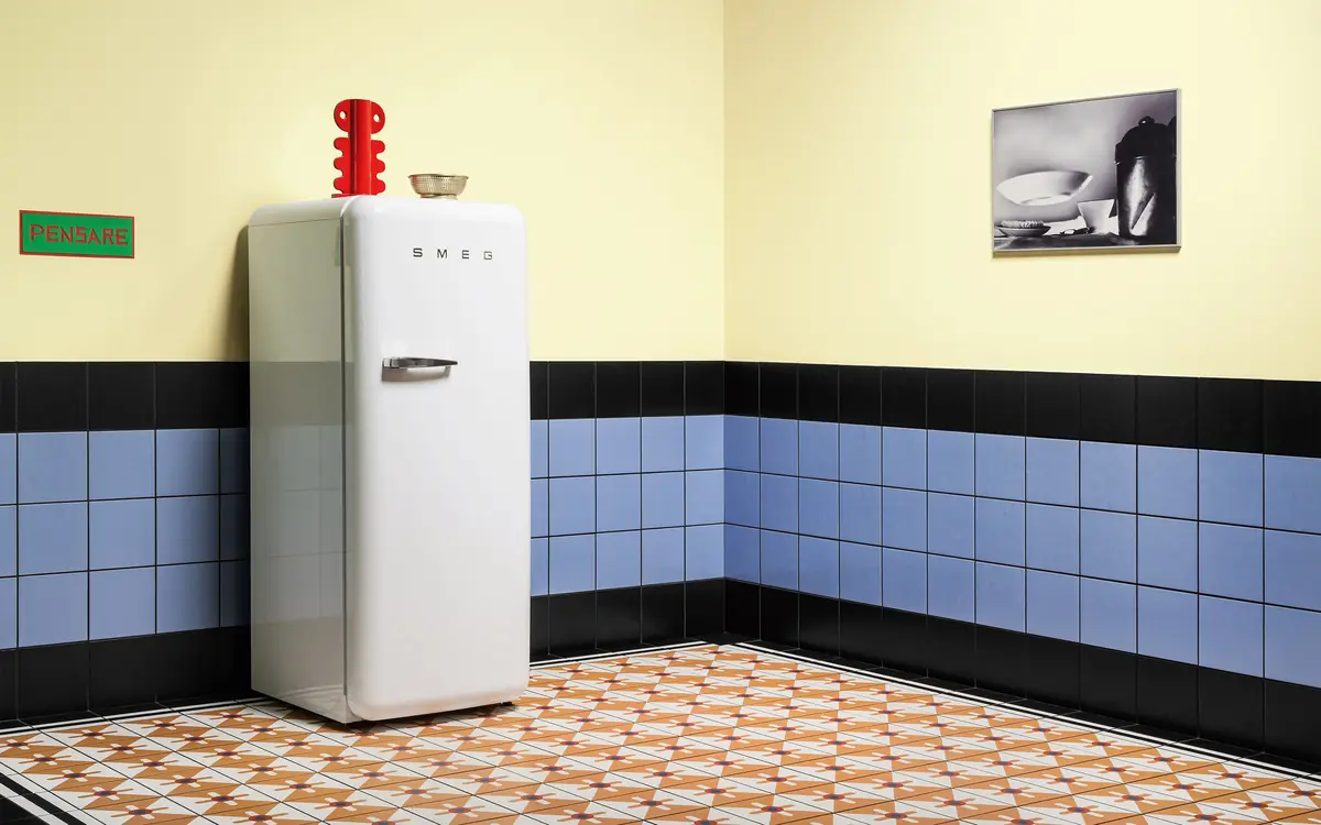 Cocina con zócalo alto de baldosas azul claro rematado en negro, pared pintada de amarillo y frigorífico blanco sobre suelo de baldosas estampadas
