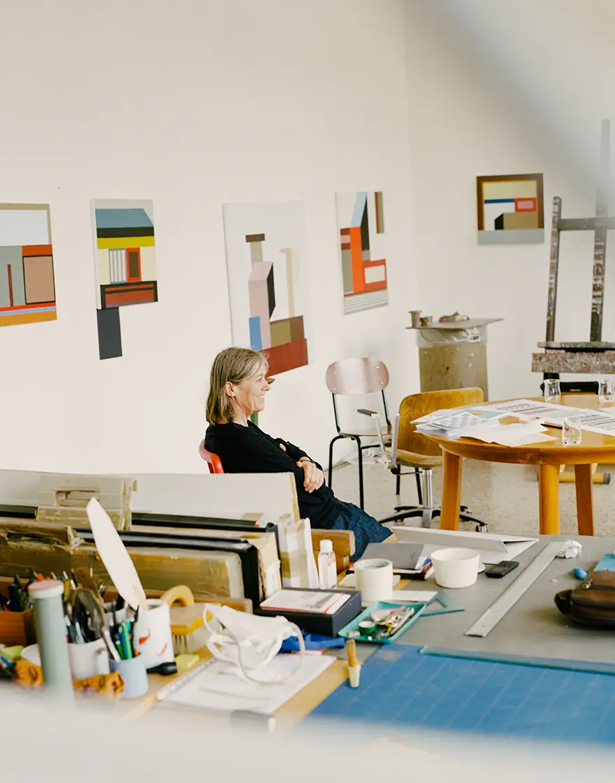 Nathalie Du Pasquier sentada en un estudio con pinturas geométricas en la pared y una mesa llena de materiales