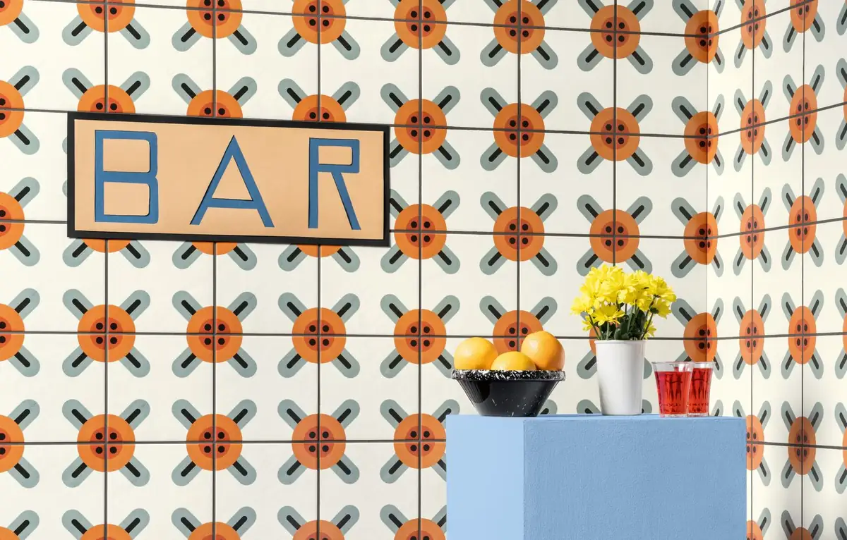 Pared revestida de baldosas blancas con flores naranjas y aspas grises, con un rótulo de bar y una peana azul con fruta y flores