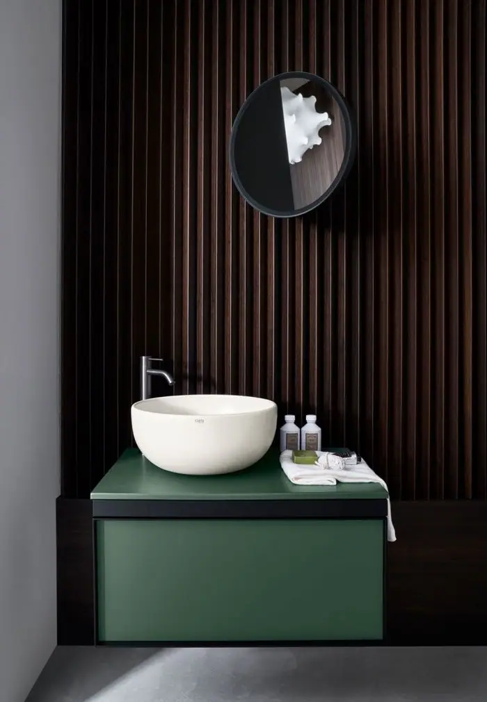 Lavabo de apoyo blanco sobre encimera verde y mueble suspendido verde con un cajón, ante una pared de listones de madera oscura