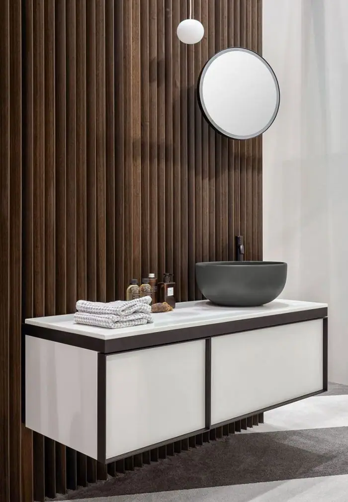 Lavabo de apoyo gris oscuro sobre encimera blanca y mueble suspendido blanco de dos cajones con marco negro, junto a una pared de listones de madera