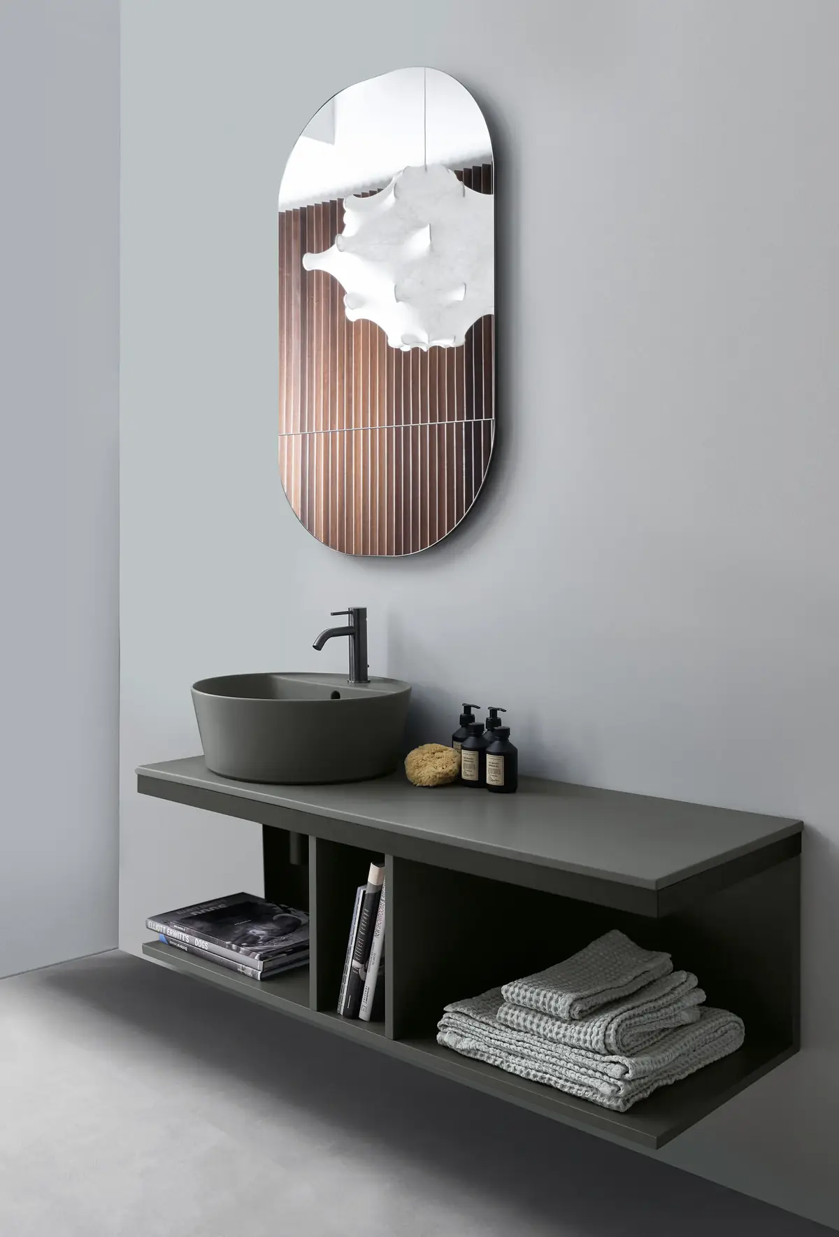 Lavabo de apoyo gris sobre encimera y mueble abierto suspendido del mismo tono, con toallas y libros en los huecos y un espejo ovalado