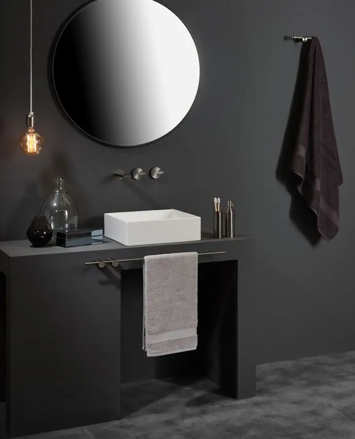 Baño oscuro con lavabo blanco sobre encimera gris, grifería mural, espejo redondo, toallero de varilla y colgador con toalla