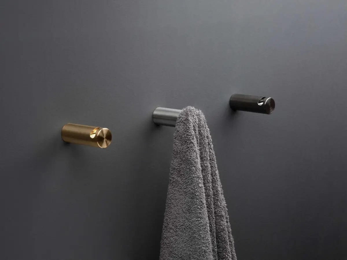 Tres colgadores cilíndricos en la pared, en dorado, acero y negro, con una toalla gris colgada del central