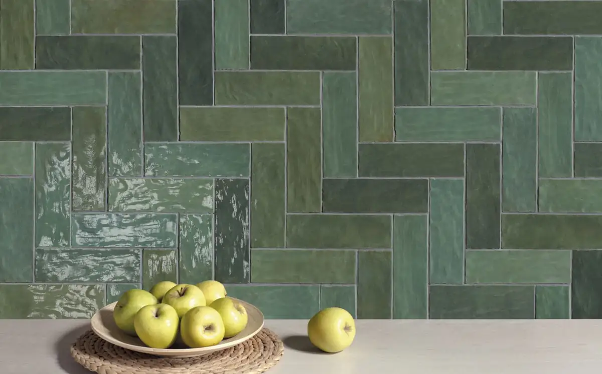 Pared revestida de piezas rectangulares verdes de esmalte irregular colocadas en espiga, con un plato de manzanas delante