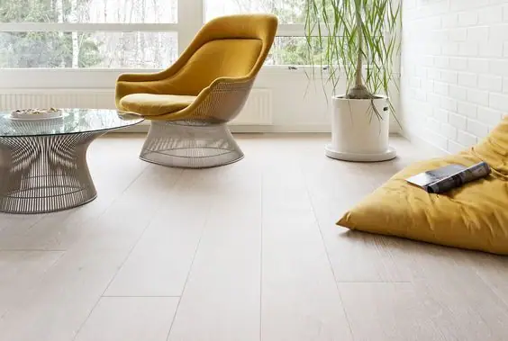Salón luminoso con butaca amarilla y mesa de cristal sobre un suelo de tablones anchos casi blancos