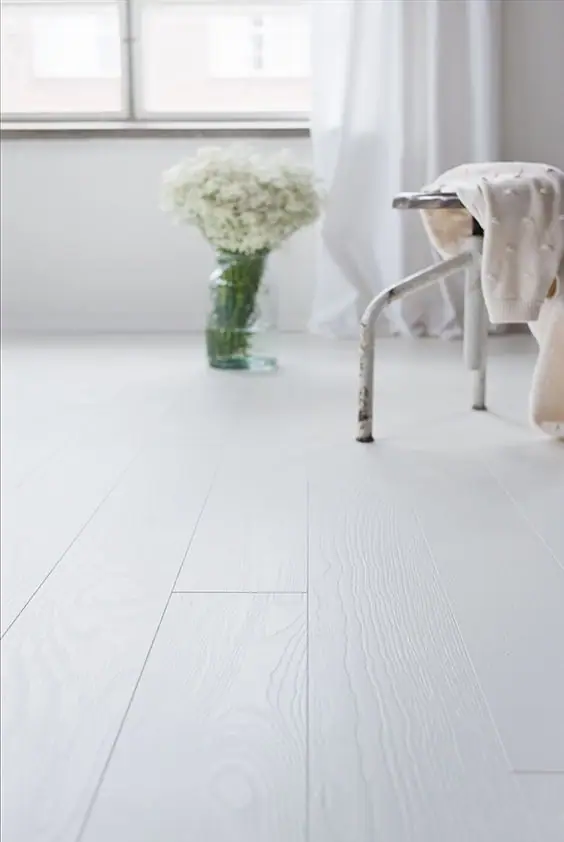 Suelo de tablones anchos blancos con veta marcada, un taburete metálico y un jarrón con flores blancas junto a una ventana
