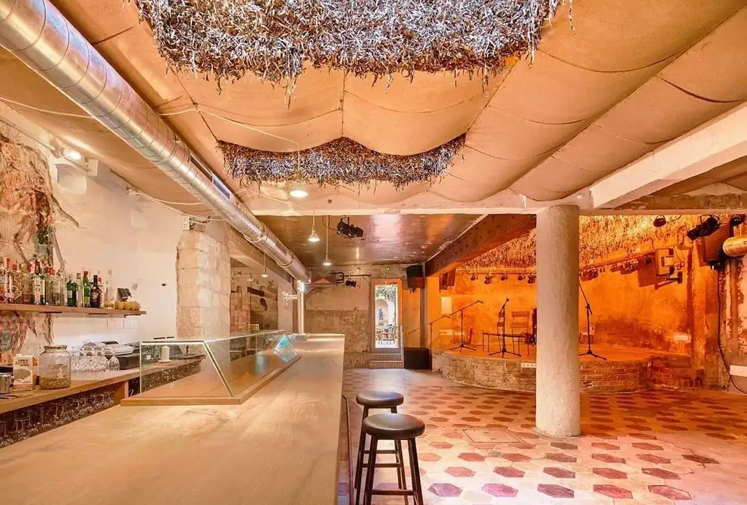 Bar con barra de madera, escenario al fondo y techo ondulado de placas de arcilla con elementos vegetales suspendidos